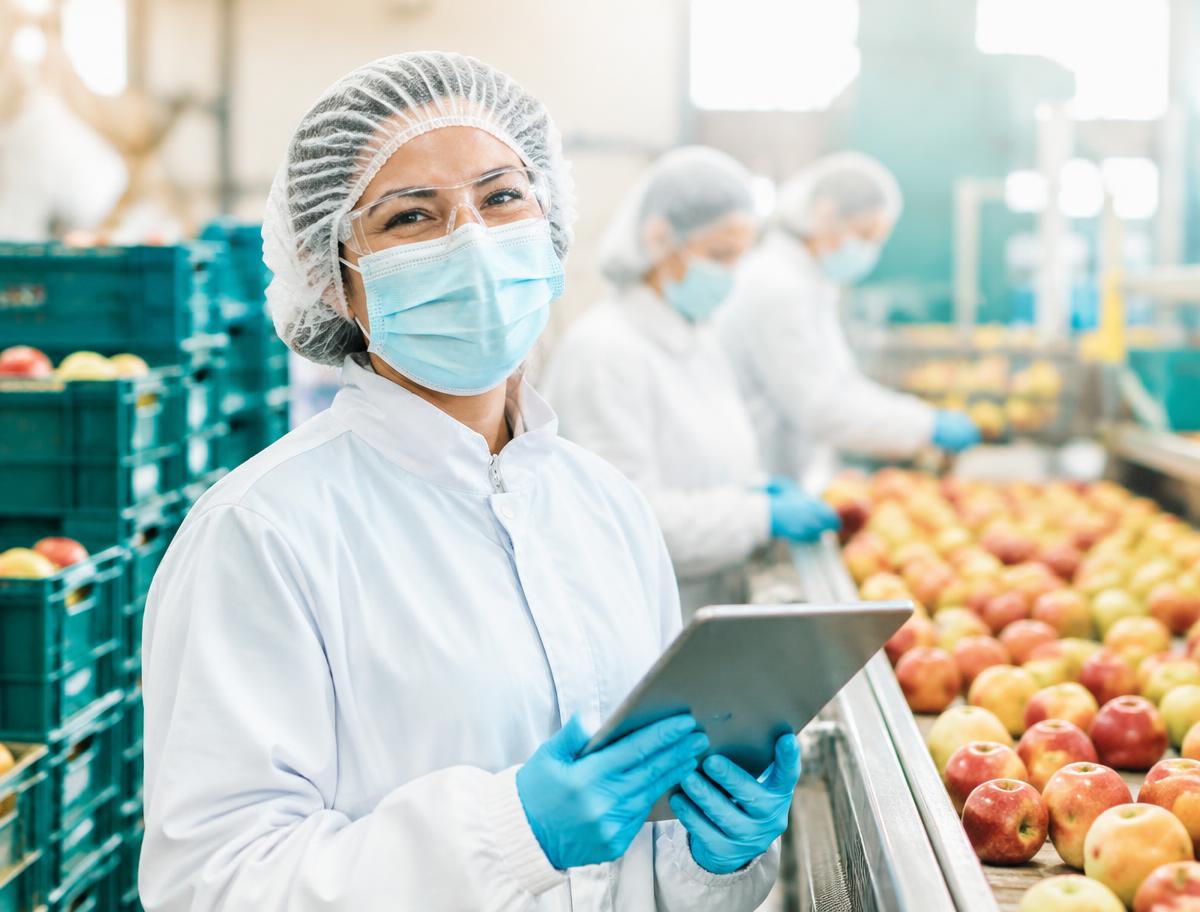 Smiling produce packing team member in full protective clothing using a tablet on the production floor