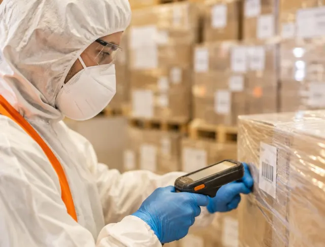 Warehouse worker in hood, mask, glasses and gloves scanning a barcode on a wrapped case