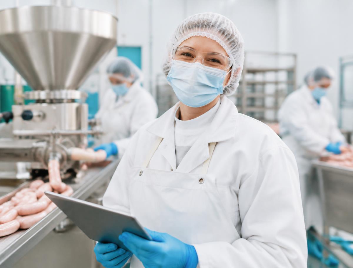 Smiling sausage & meat products team member in full protective clothing using a tablet on the production floor
