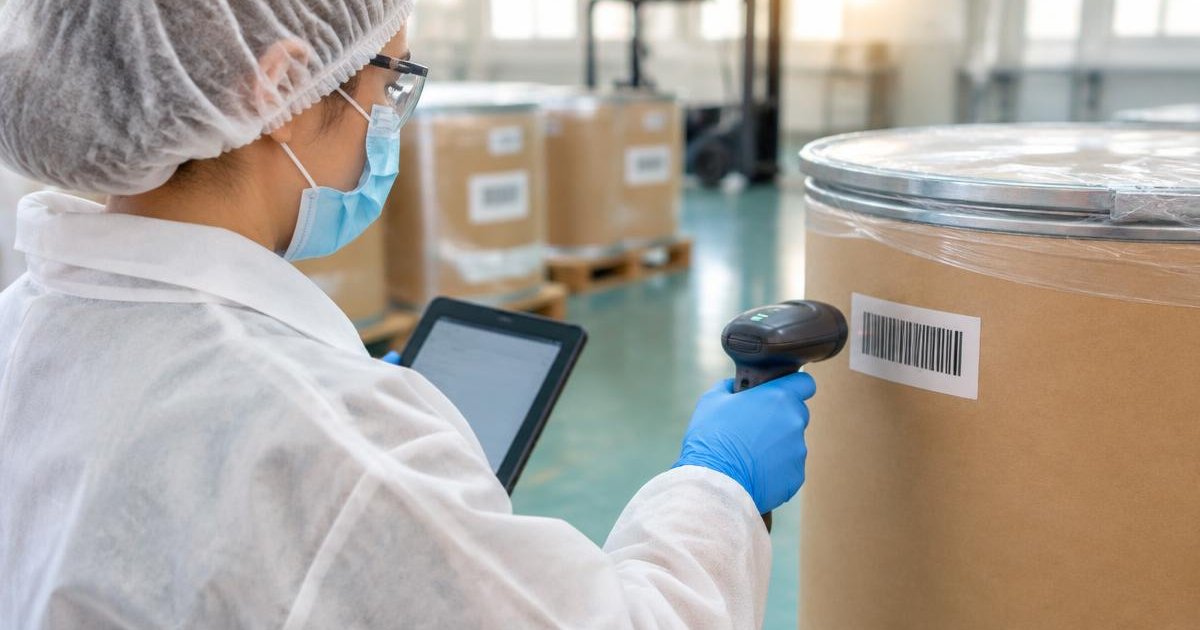 Batch ID barcode scanning: a warehouse worker in PPE aims a handheld scanner at a barcode label on a sealed ingredient drum