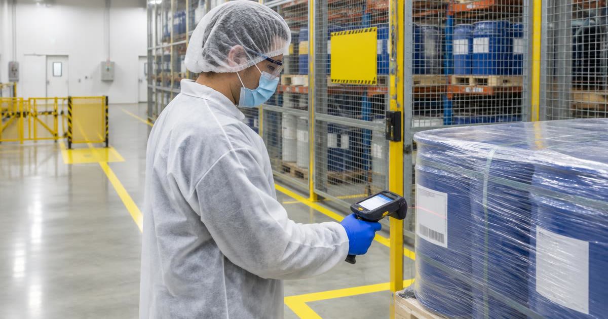 Warehouse worker in hair cover, mask and gloves scanning drums in a caged quarantine area
