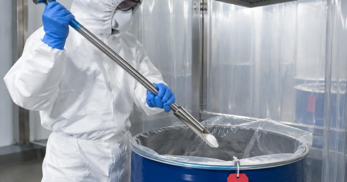 A sampler in hood, goggles, respirator and coverall draws powder from a drum with a sampling thief under a sampling booth