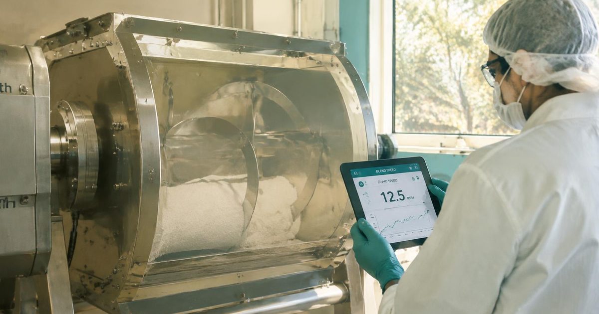 Operator in hairnet, mask, glasses and gloves monitoring blend speed on a tablet beside a gentle blender