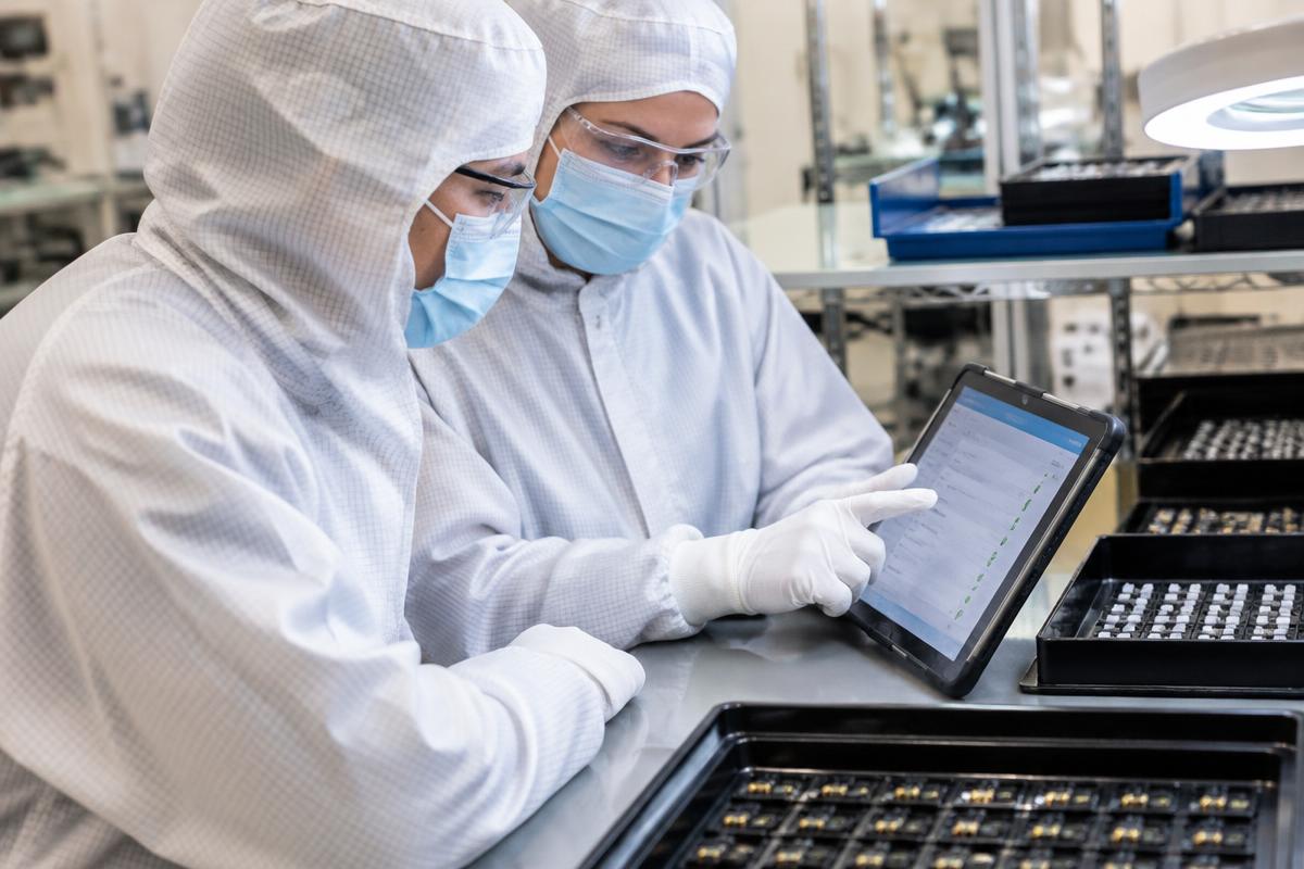 Two protected medical-device colleagues reviewing an electronic production record on a tablet at an assembly workstation